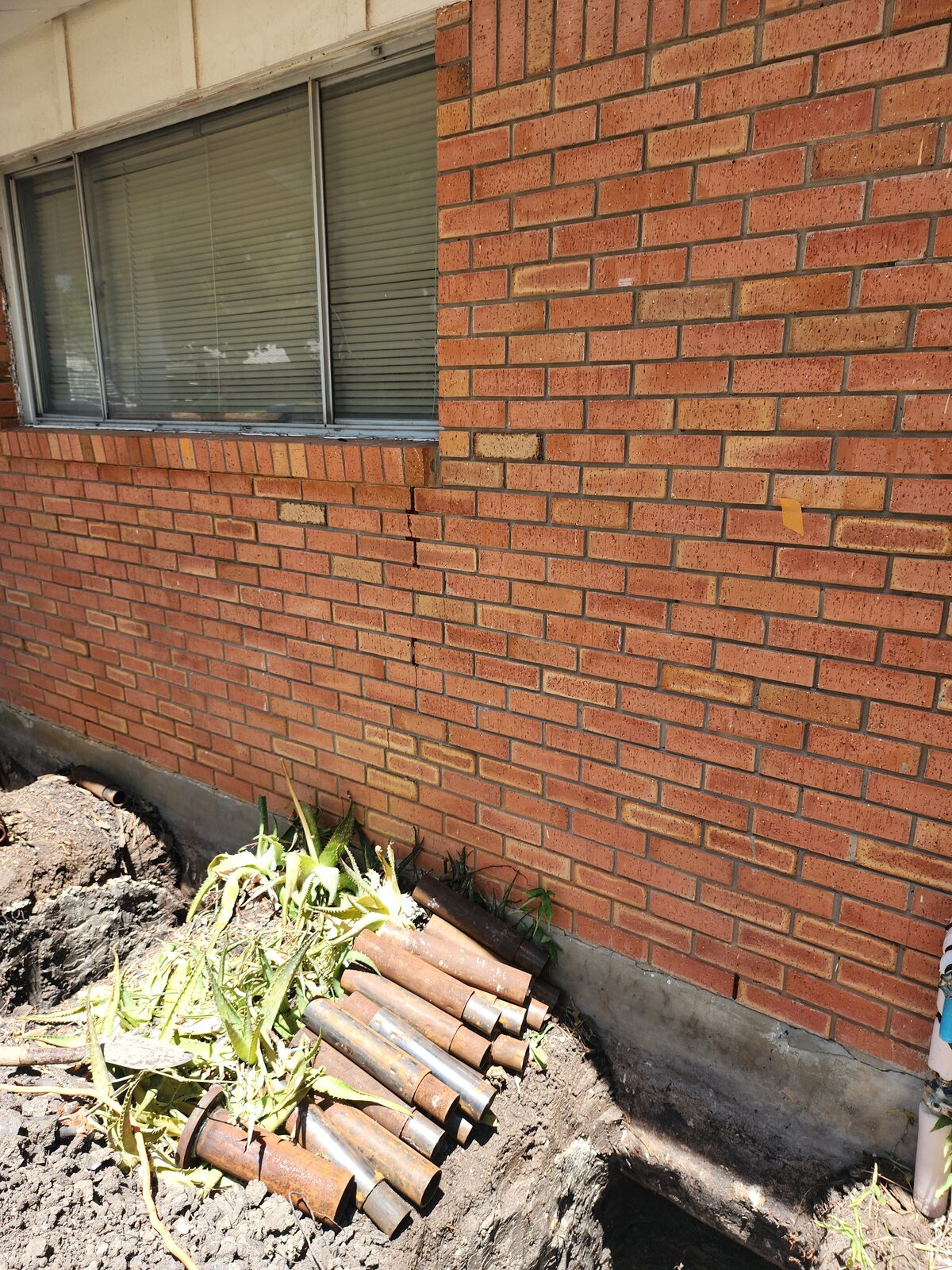 Steel pier pipe sections staged in an excavated trench beside a cracked brick wall